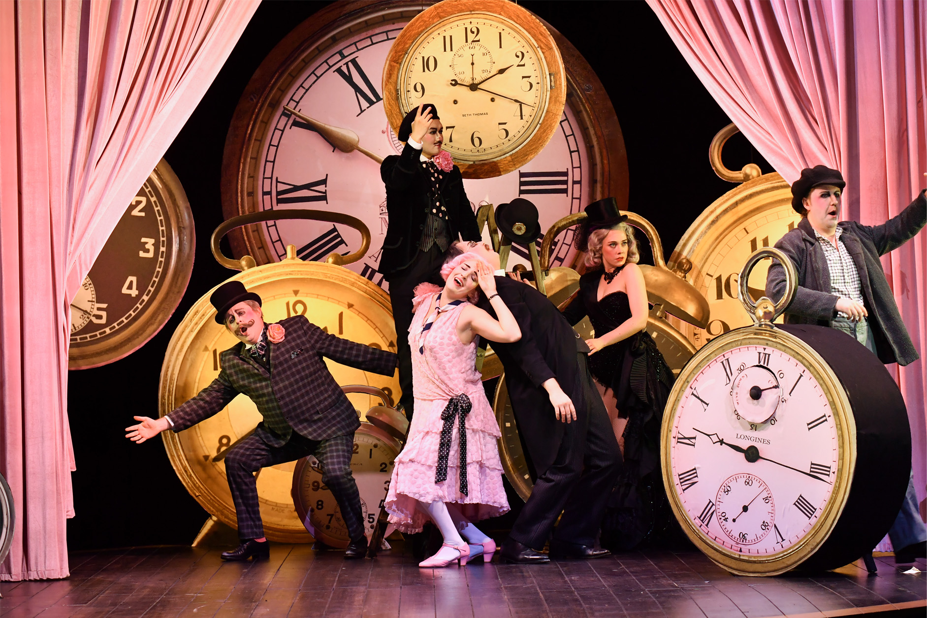 A group of opera students, wearing period costume, performing on stage, surrounded with clocks.